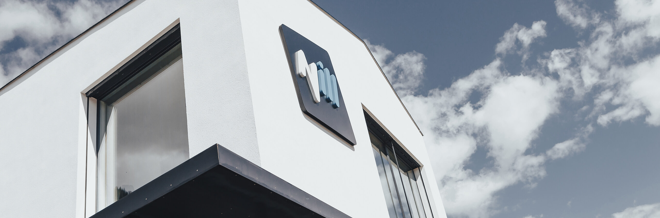 Modernes weißes Gebäude mit blau-weißem Logo, bewölkter Himmel.