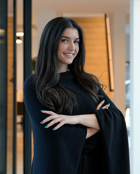 Junge Frau mit dunklen Haaren lächelt mit verschränkten Armen.