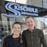 Eine Frau mit Zopf und ein Mann in dunkelgrünem Hemd stehen vor dem Schild der Skischule Bad Hofgastein
